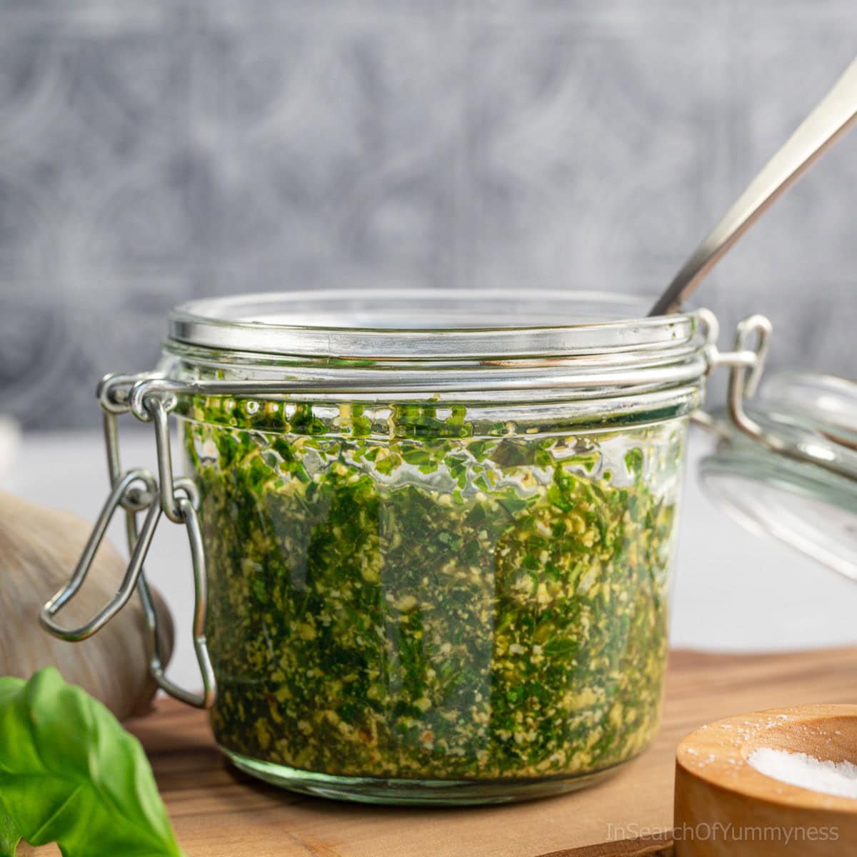 A glass jar filled with vibrant green basil pesto with walnuts, with a spoon sitting in the jar. There is a salt cellar, a sprig of fresh basil and a head of garlic around the jar.
