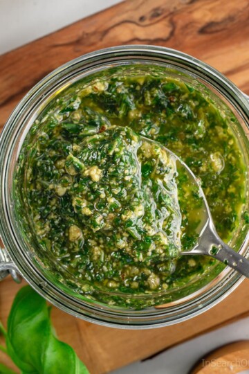 A spoonful of freshly made basil pesto with walnuts being taken out of a glass jar that is sitting on a wooden board.