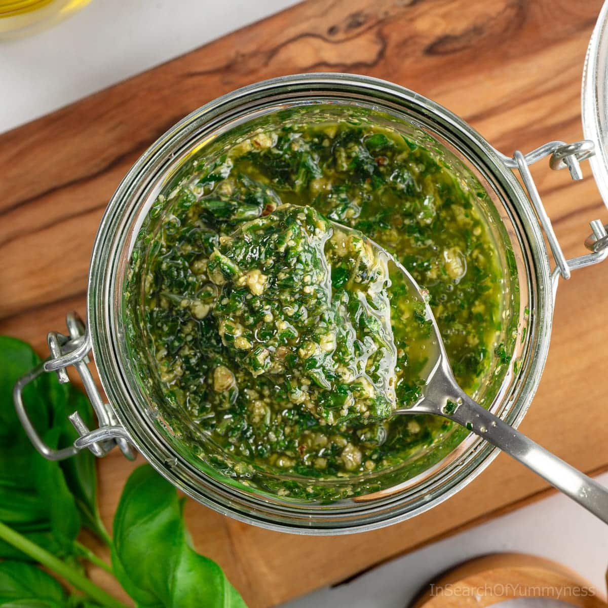A spoonful of freshly made basil pesto with walnuts being taken out of a glass jar that is sitting on a wooden board.