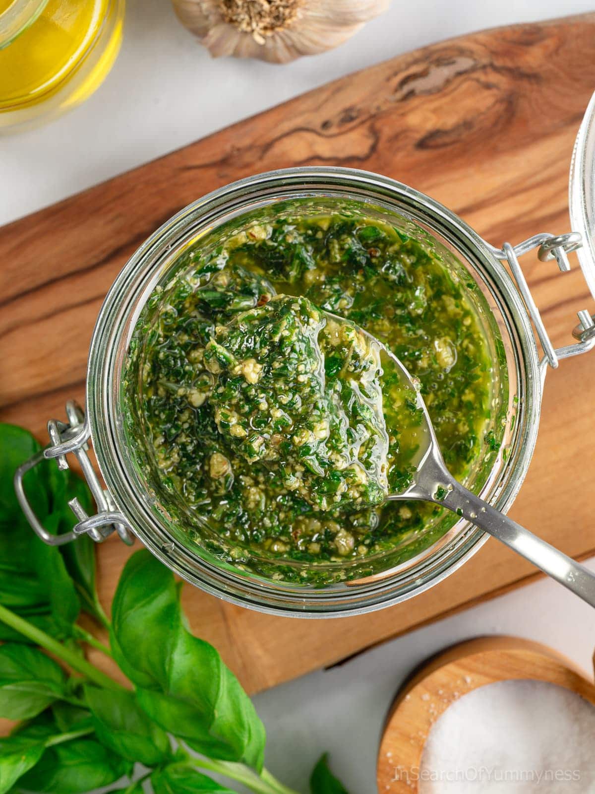 A spoonful of freshly made basil pesto with walnuts being taken out of a glass jar that is sitting on a wooden board. There's a salt cellar, a sprig of fresh basil, and a head of garlic around the jar.
