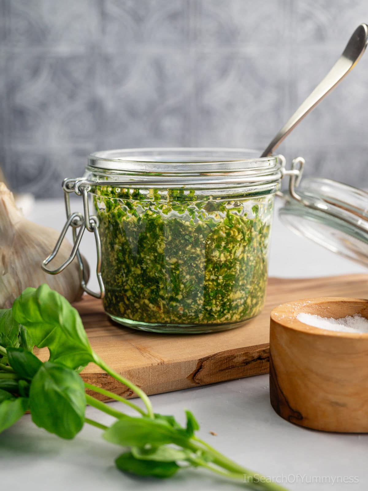 A glass jar filled with vibrant green basil pesto with walnuts, with a spoon sitting in the jar. There is a salt cellar, a sprig of fresh basil and a head of garlic around the jar.