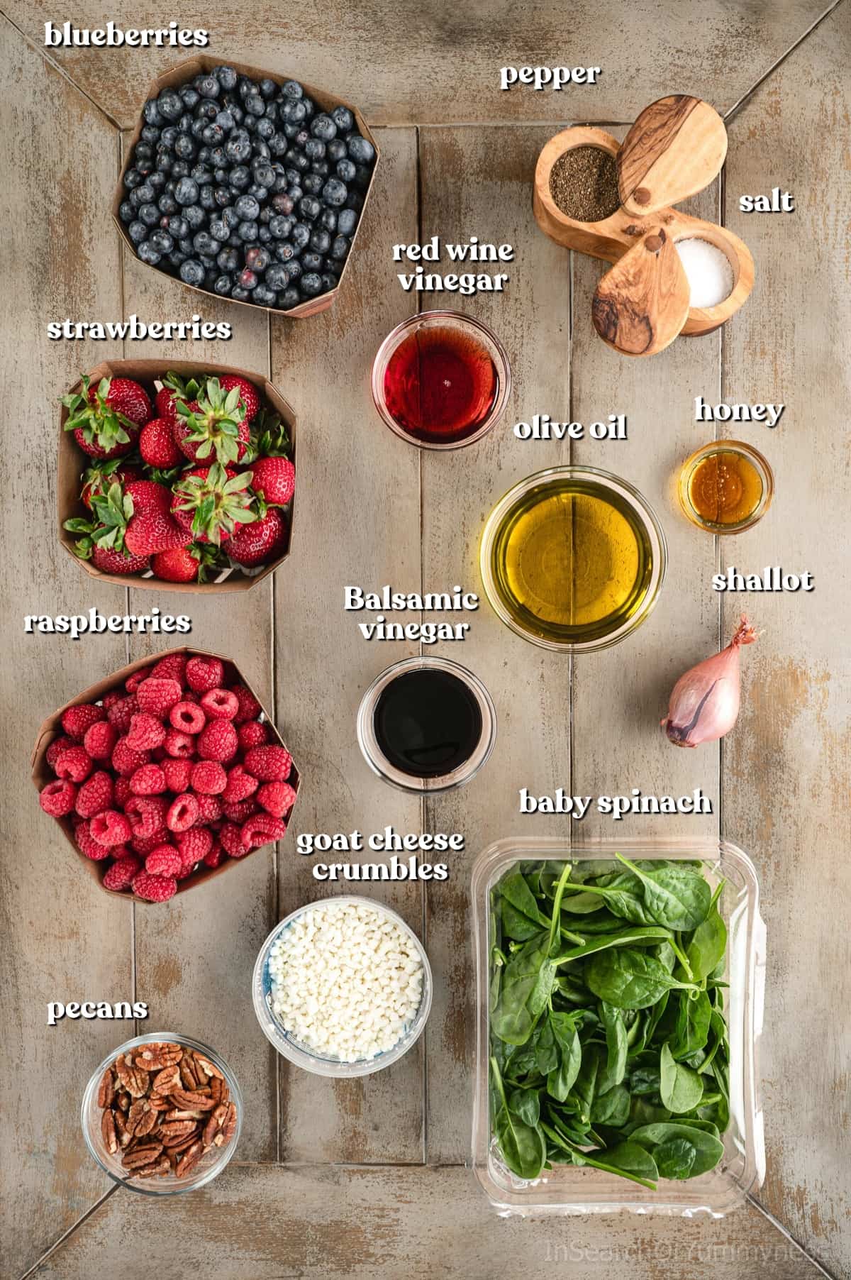 Ingredients needed to make this berry spinach salad recipe in little bowls on a light wooden table. A large container of baby spinach is sitting next to the bowls and three berry boxes with the blueberries, strawberries and raspberries.