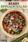 A wooden bowl full of baby spinach, fresh berries, goat cheese crumbles and toasted pecans, next to a glass jug of raspberry vinaigrette, a pepper mill, a can of forks and wooden salad servers. Text overlay says "Berry Spinach Salad".