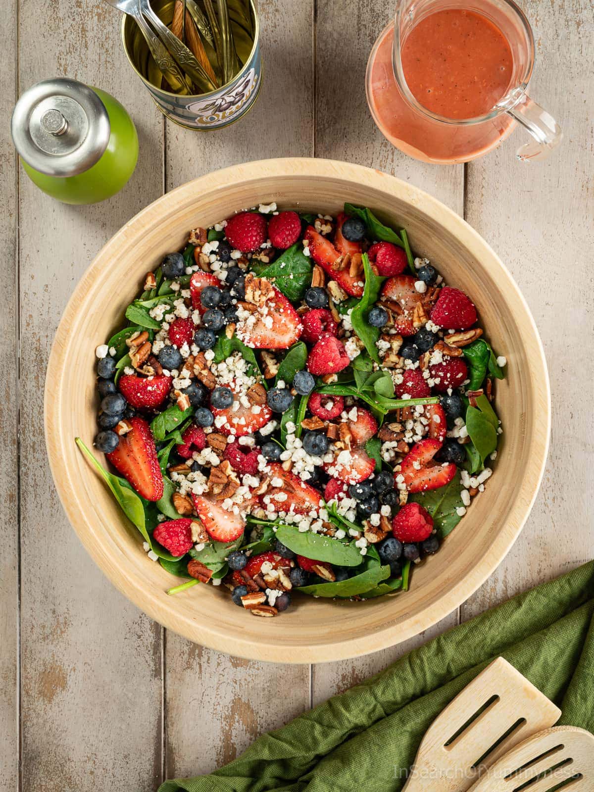 A wooden bowl full of baby spinach, fresh berries, goat cheese crumbles and toasted pecans, next to a glass jug of raspberry vinaigrette, a pepper mill, a can of forks and wooden salad servers.