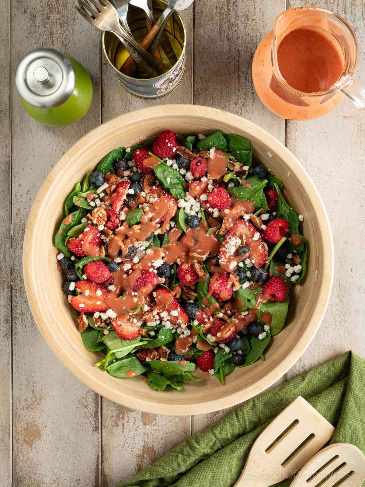A wooden bowl full of baby spinach, fresh berries, goat cheese crumbles and toasted pecans, dressed with raspberry vinaigrette, next to a glass jug of extra vinaigrette, a pepper mill, a can of forks and wooden salad servers.