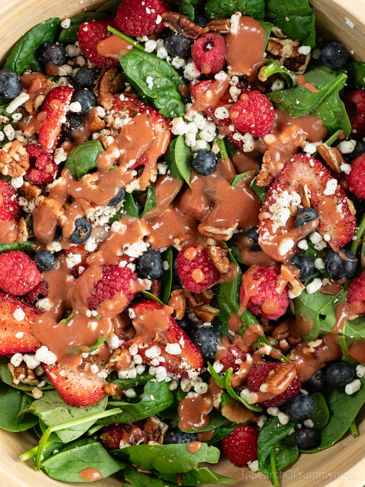 A close up of a wooden bowl full of baby spinach, fresh berries, goat cheese crumbles and toasted pecans, dressed in a raspberry vinaigrette.
