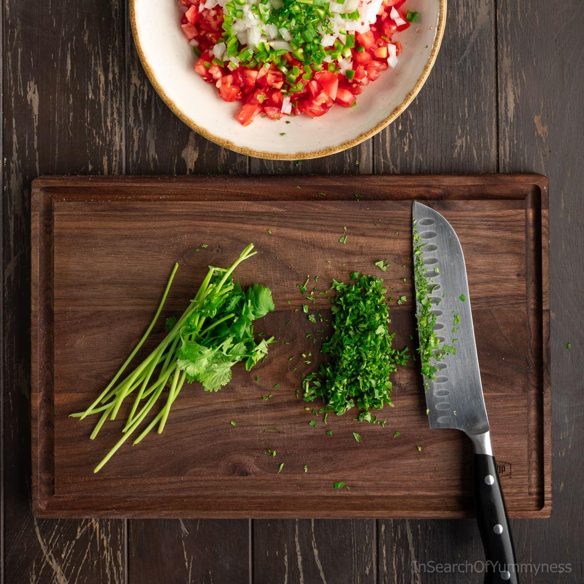 A dark walnut cutting board with finely chopped fresh cilantro and a santoku knife. There's a large serving bowl with diced tomatoes, onion, and serrano peppers above the board.