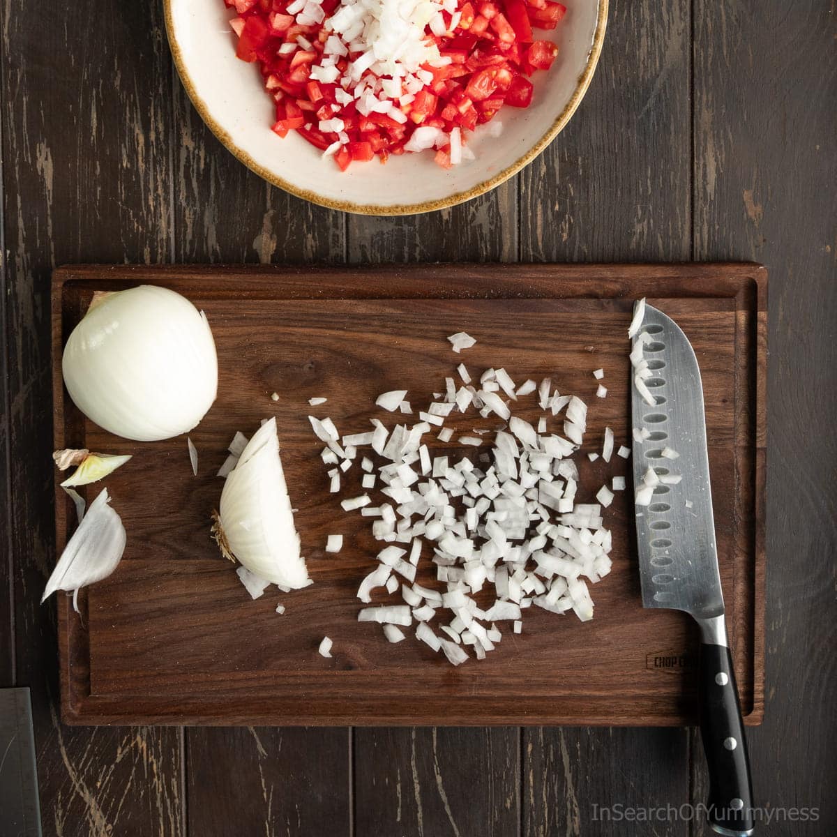 A dark walnut cutting board with finely diced white onion and a santoku knife. There's a large serving bowl with diced tomatoes and onion above the board.