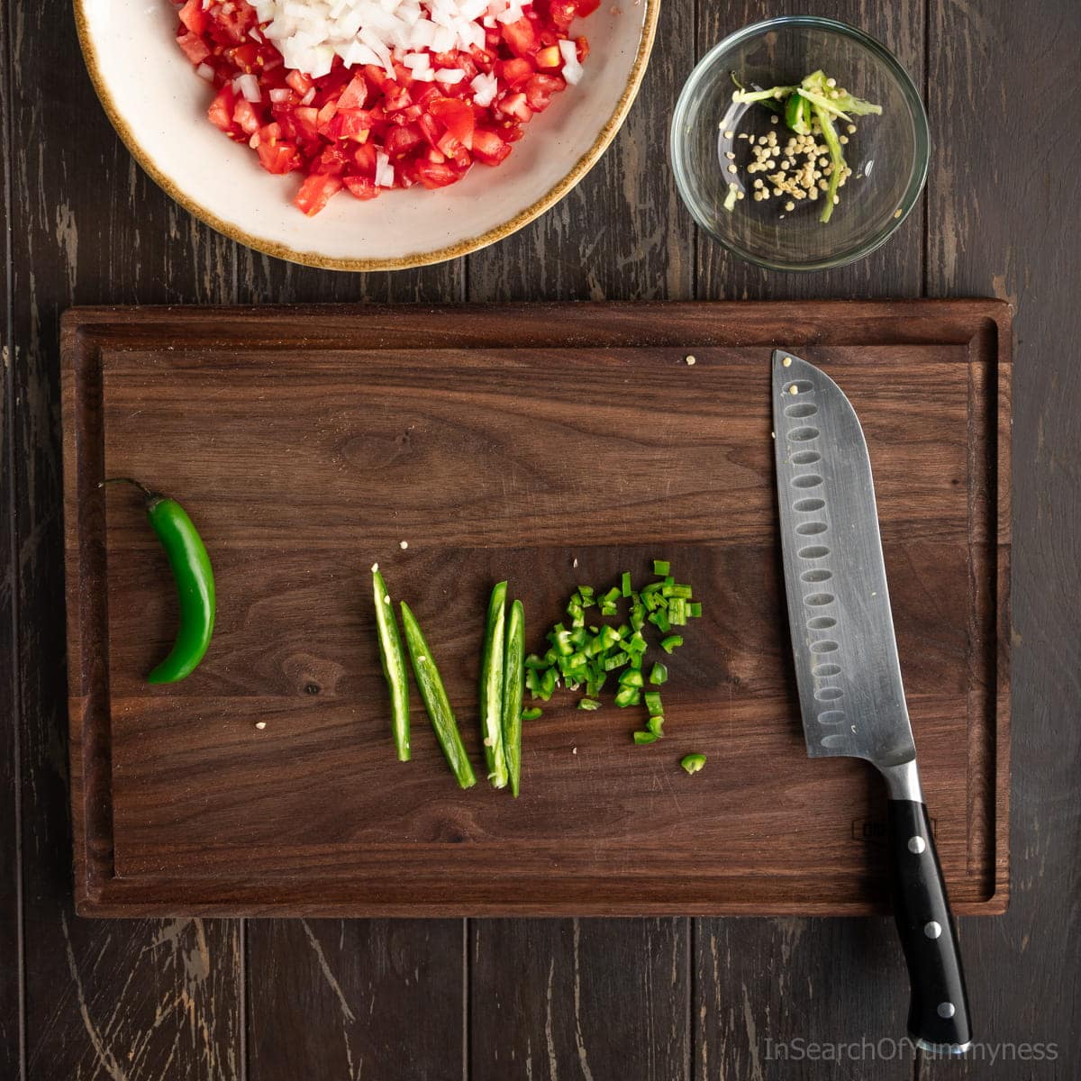 A dark walnut cutting board with finely diced serrano peppers and a santoku knife. There's a small clear bowl with a discarded serrano seeds and inner ribs above the board.