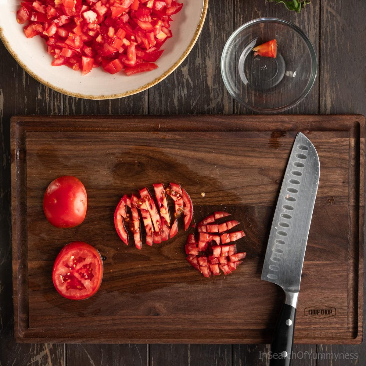 A dark walnut cutting board with finely diced tomatoes and a santoku knife. There's a small clear bowl with a discarded tomato core above the board.