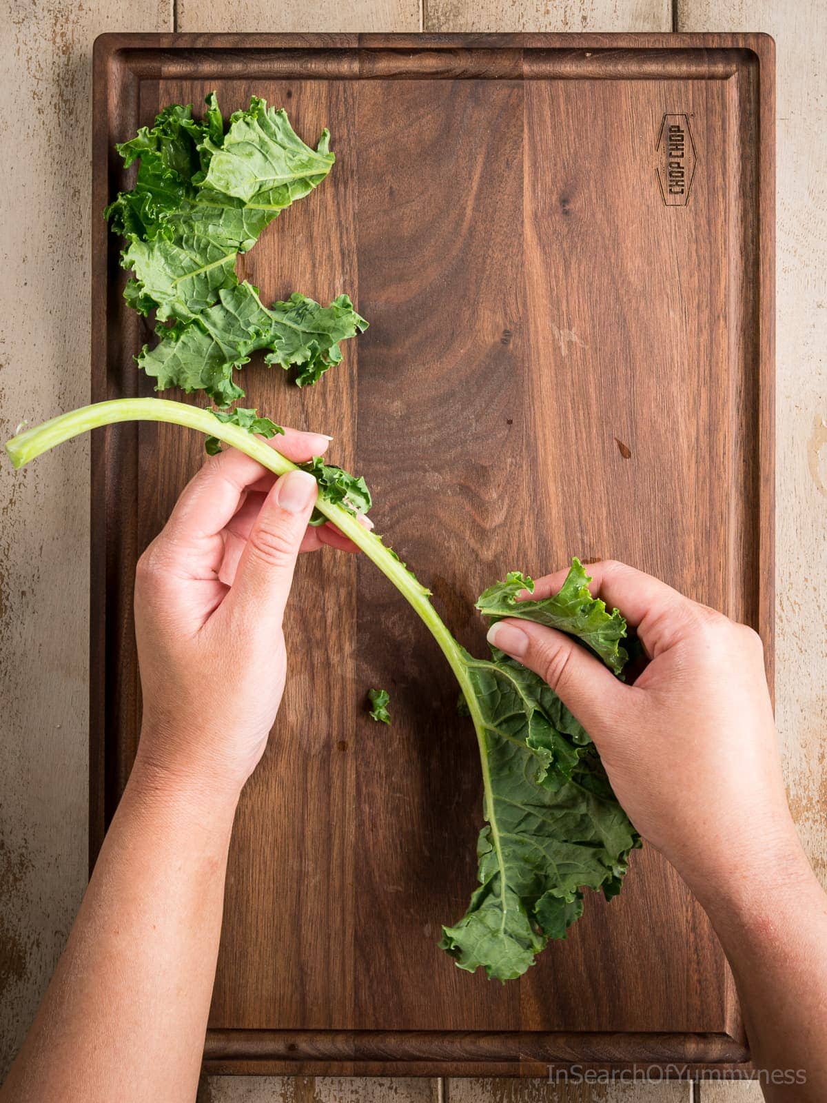 Someone demonstrating how to remove a kale leaf from the stem over a brown cutting board.