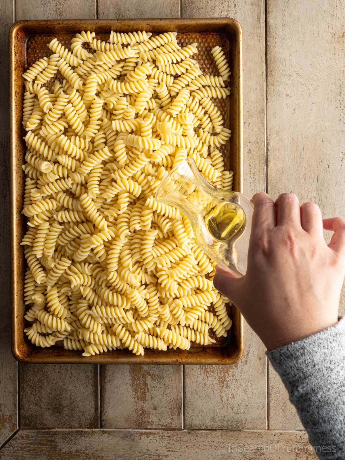 Someone adding olive oil to a baking sheet full of freshly cooked hot pasta.