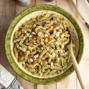 A large bowl filled with freshly made pesto pasta salad with feta, garnished with olives, marinated artichoke hearts, and sun-dried tomatoes. There is a wooden spoon in the bowl.