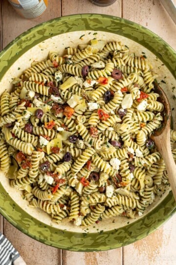A large bowl filled with freshly made pesto pasta salad with feta, garnished with olives, marinated artichoke hearts, and sun-dried tomatoes. There is a wooden spoon in the bowl.