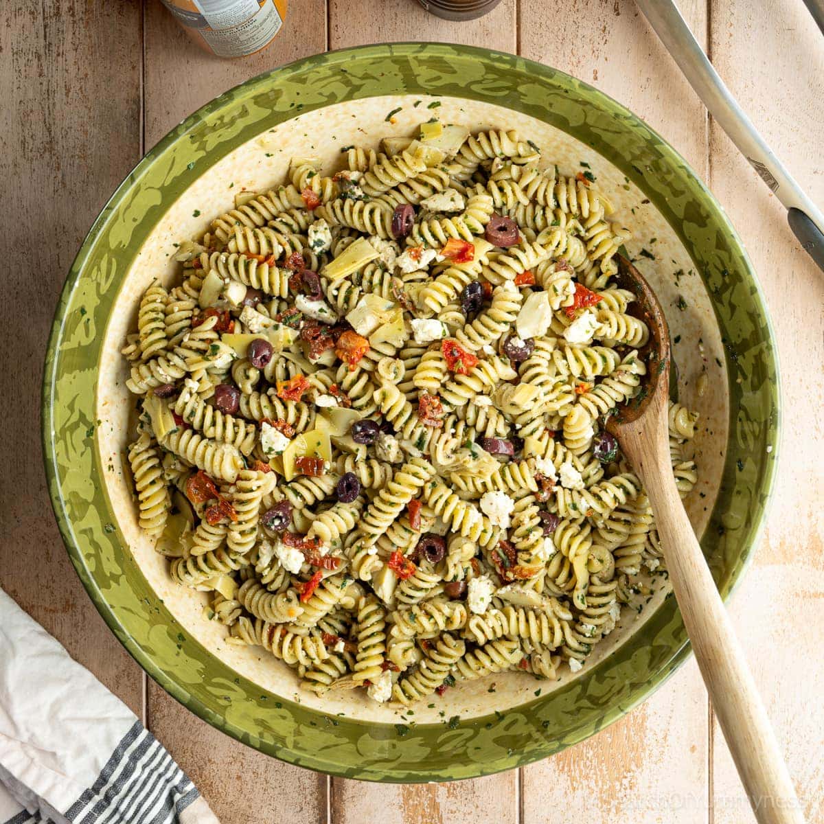 A large bowl filled with freshly made pesto pasta salad with feta, garnished with olives, marinated artichoke hearts, and sun-dried tomatoes. There is a wooden spoon in the bowl.