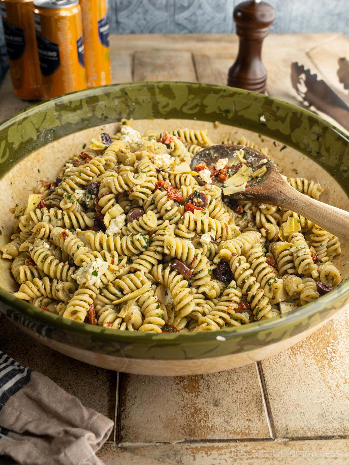 A large bowl filled with freshly made pesto pasta salad with feta, garnished with olives, marinated artichoke hearts, and sun-dried tomatoes. There is a wooden spoon in the bowl.