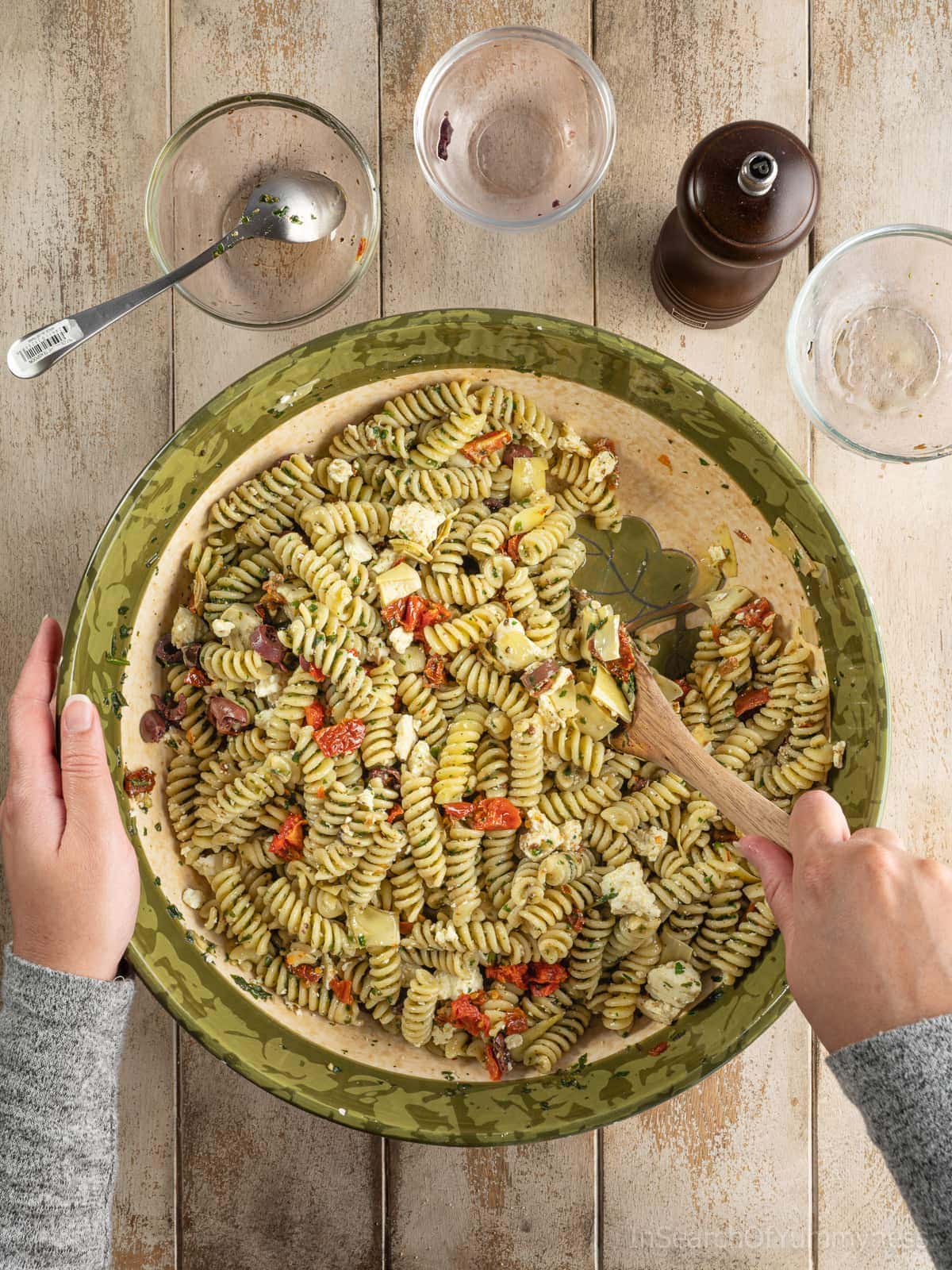 Someone using a wooden spoon to stir pesto pasta salad with feta, garnished with olives, marinated artichoke hearts, and sun-dried tomatoes.