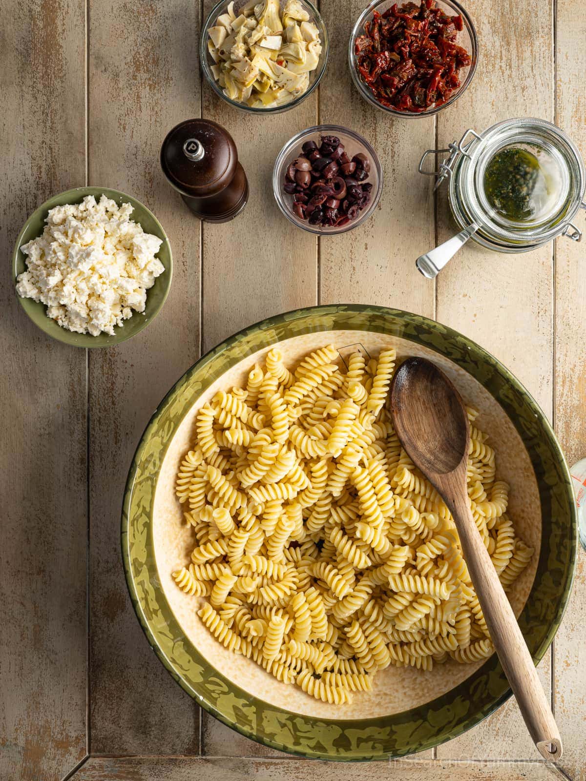 A large bowl filled with freshly cooked pasta, with a wooden spoon sitting in it. There are bowls of garnishes around the large bowl with olives, marinated artichoke hearts, and sun-dried tomatoes.