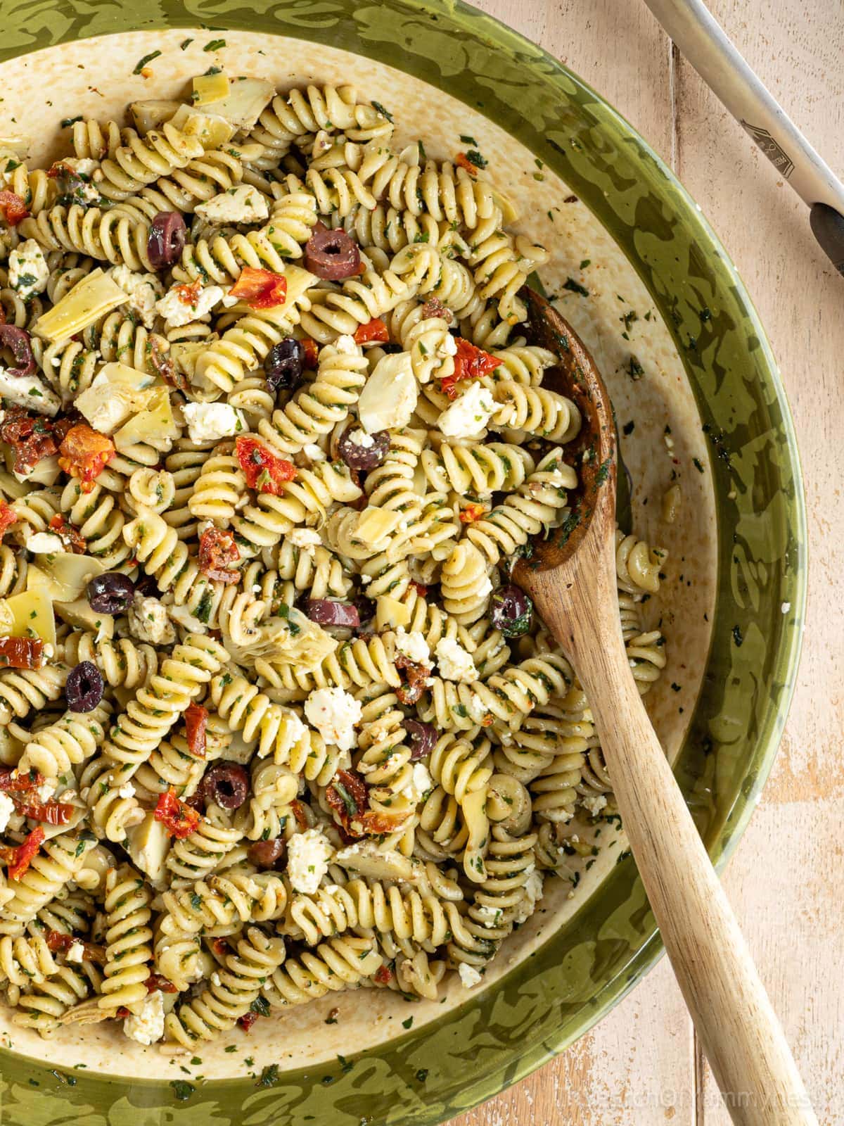 A large bowl filled with freshly made pesto pasta salad with feta, garnished with olives, marinated artichoke hearts, and sun-dried tomatoes. There is a wooden spoon in the bowl.