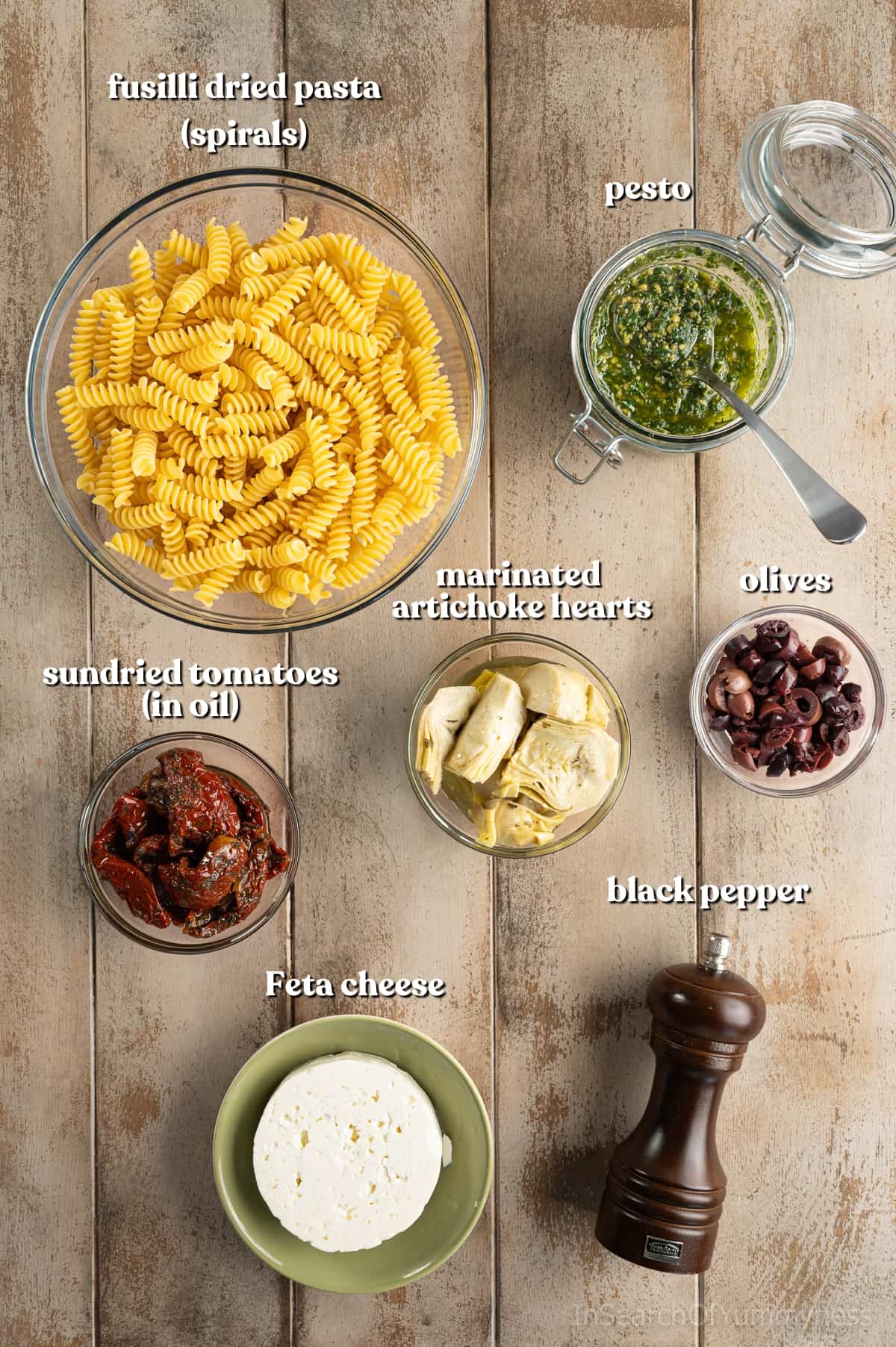 Ingredients for homemade pesto pasta salad with feta, fusilli, pesto, olives, marinated artichoke hearts, sun-dried tomatoes, and black pepper on a light wooden board.