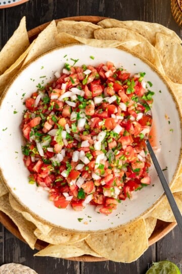 A large white bowl of pico de gallo fresh salsa surrounded by tortilla chips. There is a spoon in the bowl, and a glass next to it.