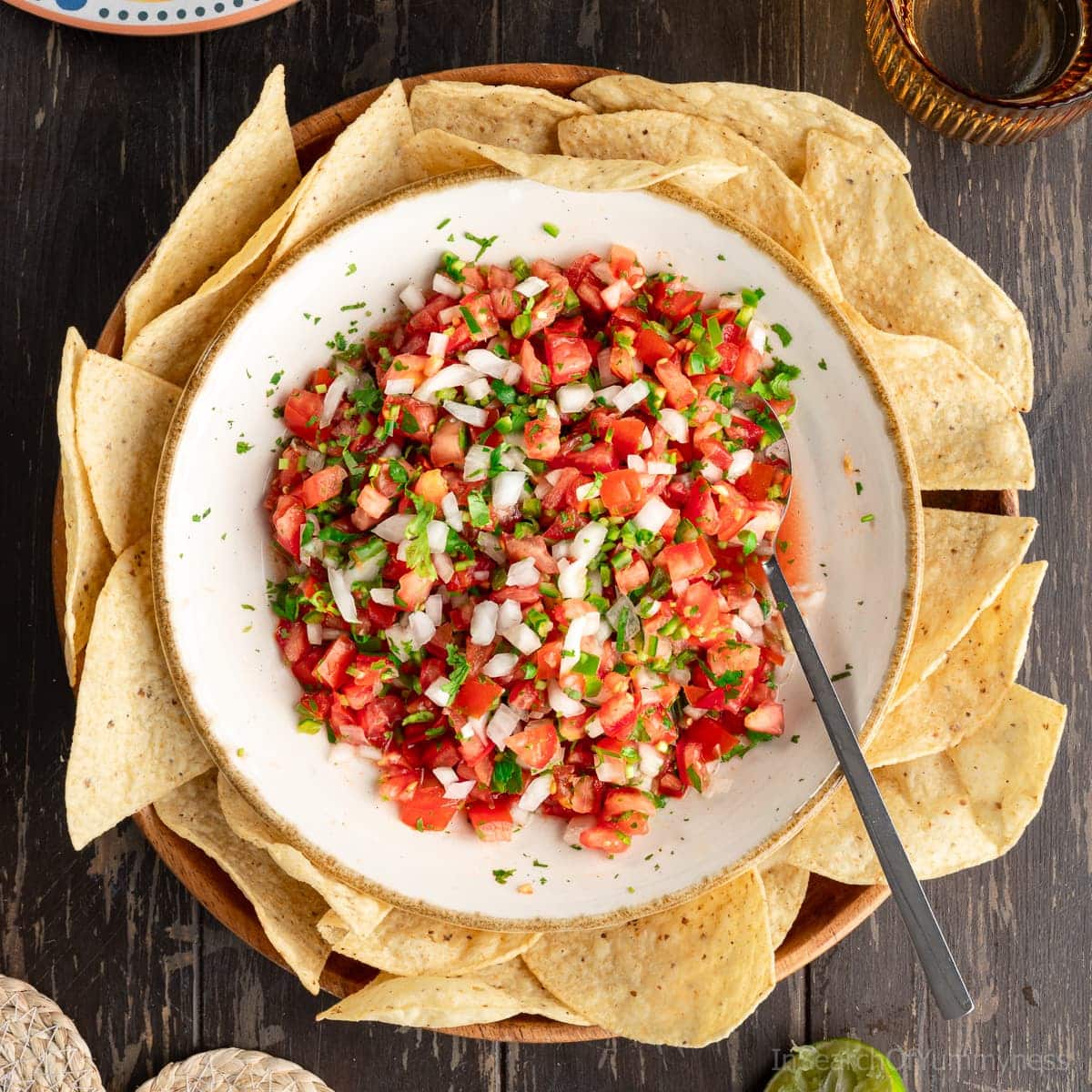 A large white bowl of pico de gallo fresh salsa surrounded by tortilla chips. There is a spoon in the bowl, and a glass next to it.