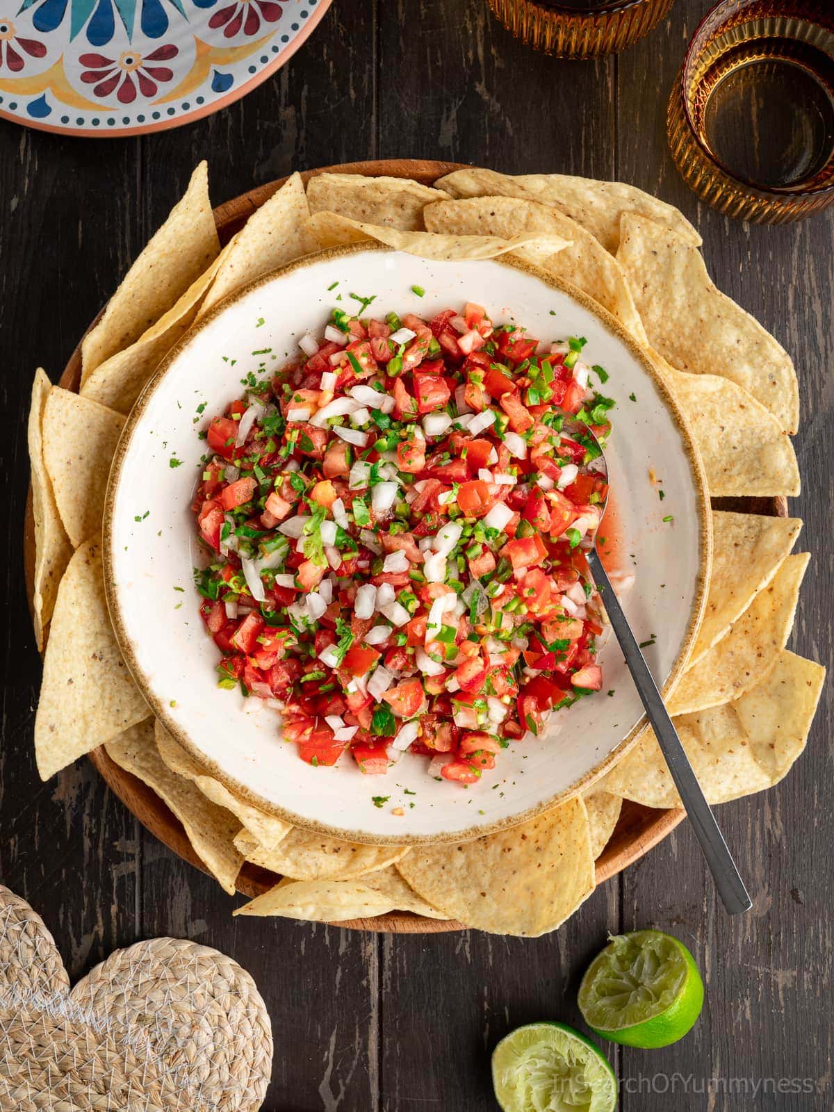A large bowl with finely diced tomatoes, onion, serrano peppers and chopped cilantro. There is a spoon in the bowl, and the bowl has tortilla chips around it.