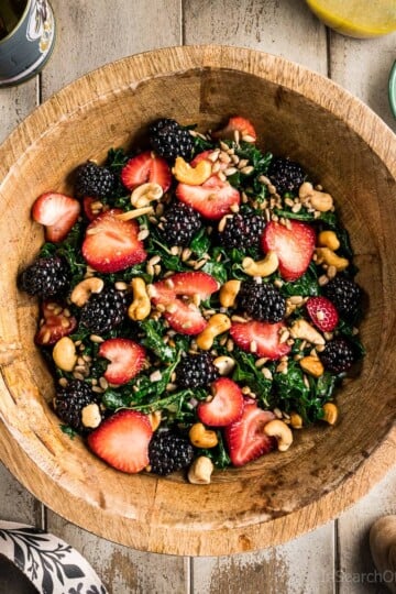 A large wooden bowl full of summer kale salad with strawberries, blackberries, sunflower seeds and cashews.
