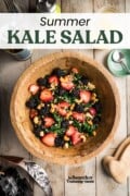 A large wooden bowl full of summer kale salad with strawberries, blackberries, sunflower seeds and cashews, sitting on a picnic table. Text overlay says "Summer Kale Salad".