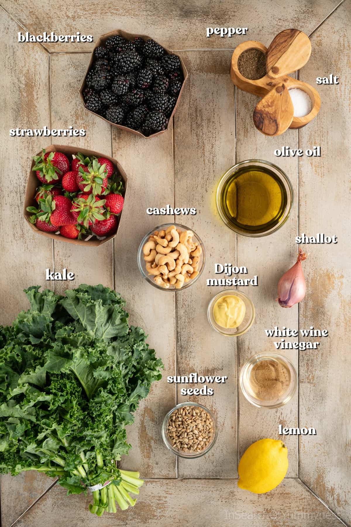 Ingredients needed to make this summer kale salad recipe in little bowls on a light wooden table. A large bunch of kale is sitting next to the bowls and two berry boxes with the blackberries and raspberries.