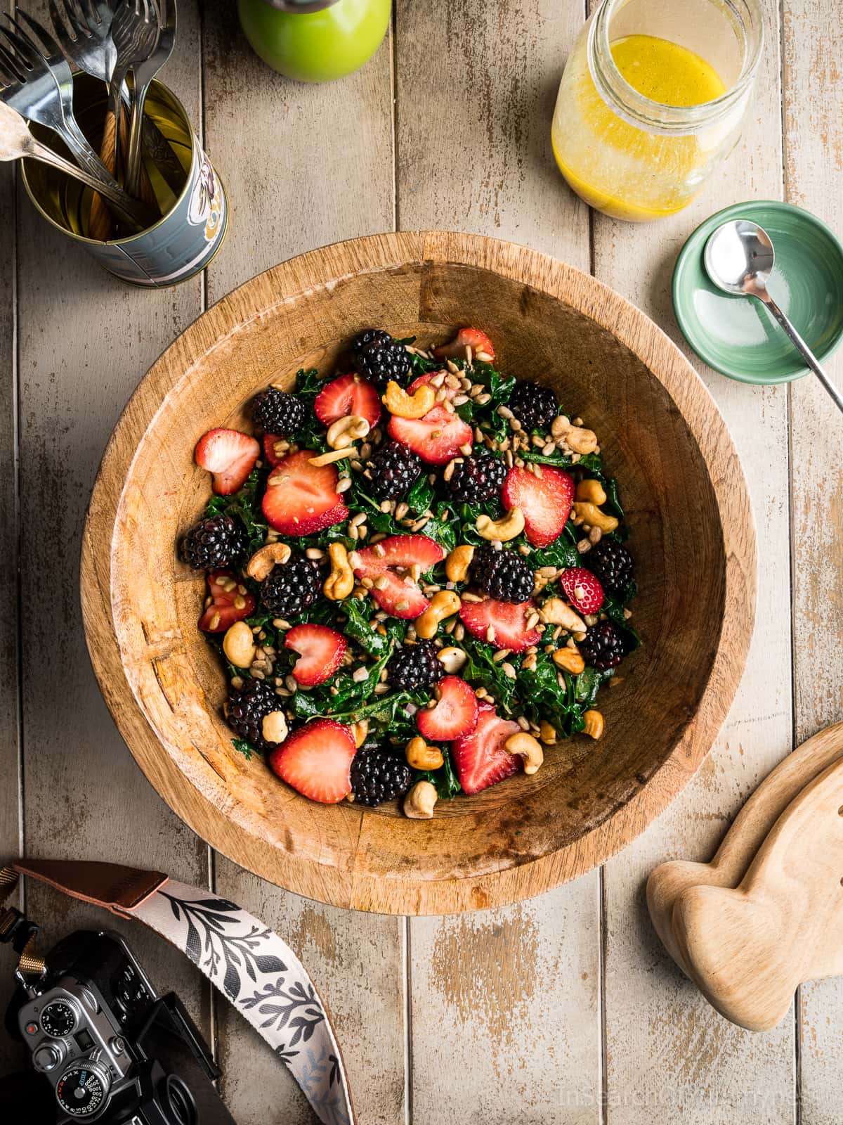 A large wooden bowl full of summer kale salad with strawberries, blackberries, sunflower seeds and cashews. There is a mason jar with bright yellow honey Dijon dressing next to the bowl, as well as a camera, pepper mill, and a tin with forks.