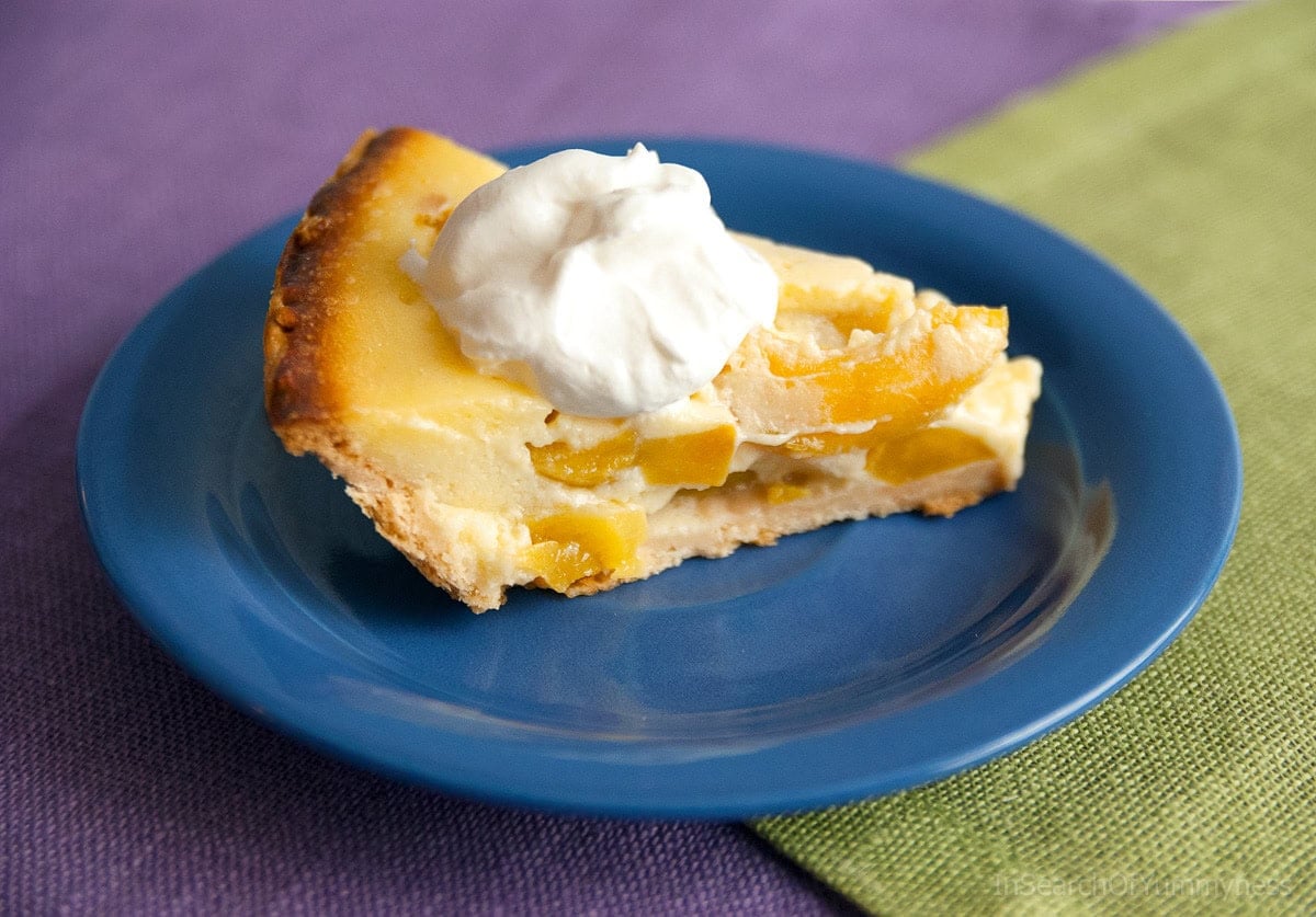 A side view of a slice of freshly baked sour cream peach pie topped with a dollop of whipped cream. The pie is on a blue plate, on a green tablecloth.