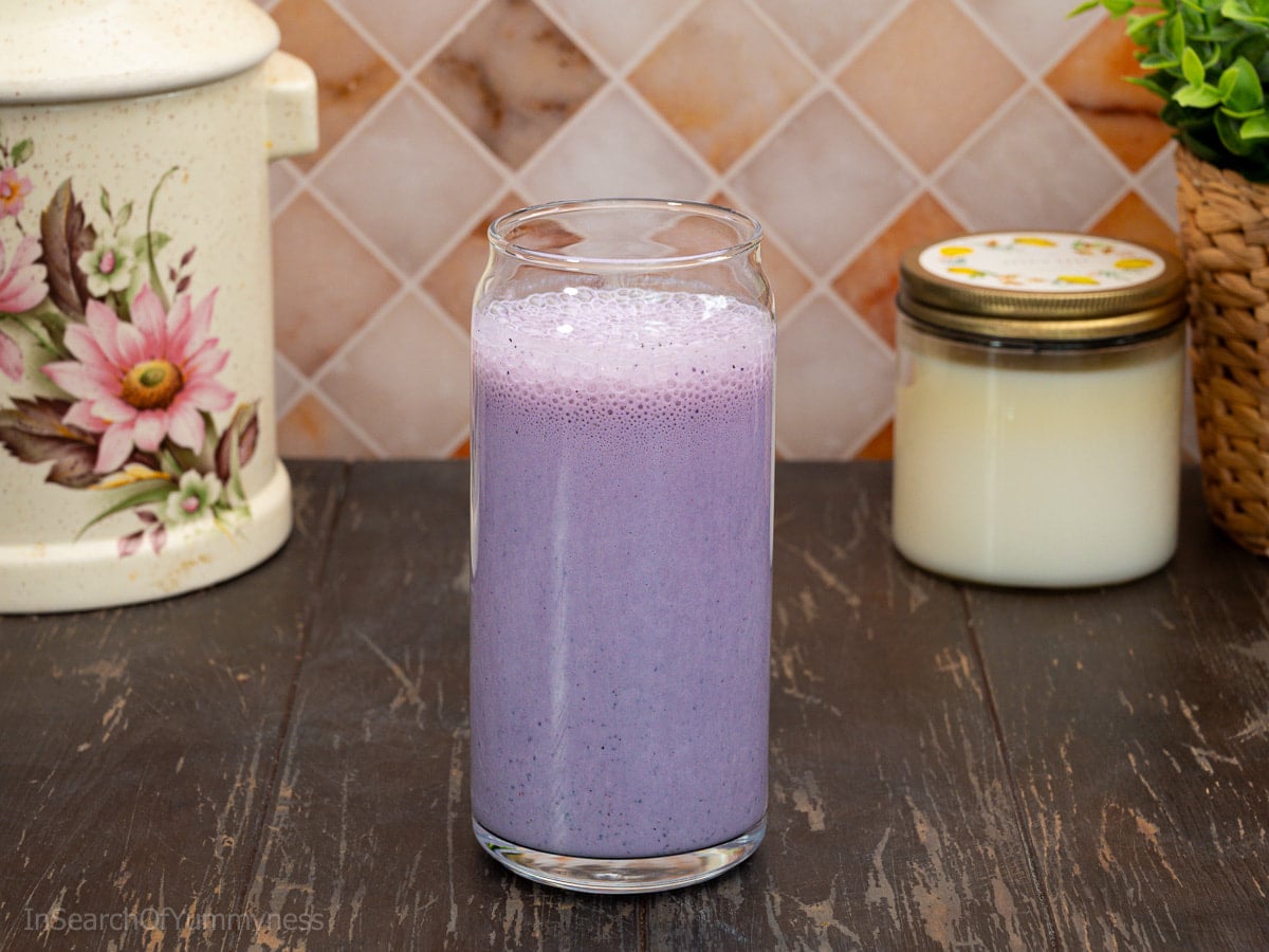 A tall glass filled with frothy purple blueberry oat smoothie. The glass is sitting on a wooden table next to a candle, a plant, and a large ceramic jar.