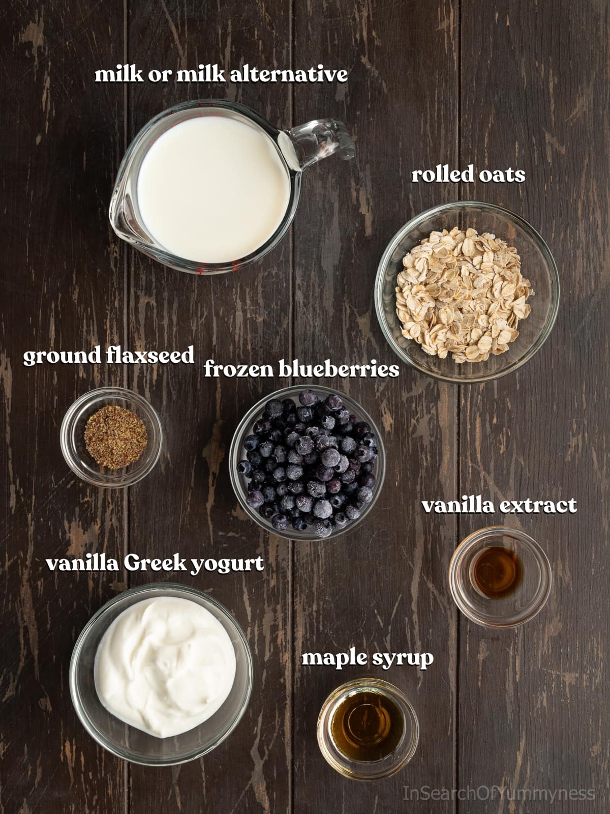 Ingredients needed to make blueberry oat smoothies including milk, oats, ground flaxseed, blueberries, vanilla, maple syrup and Greek yogurt on a wooden board.