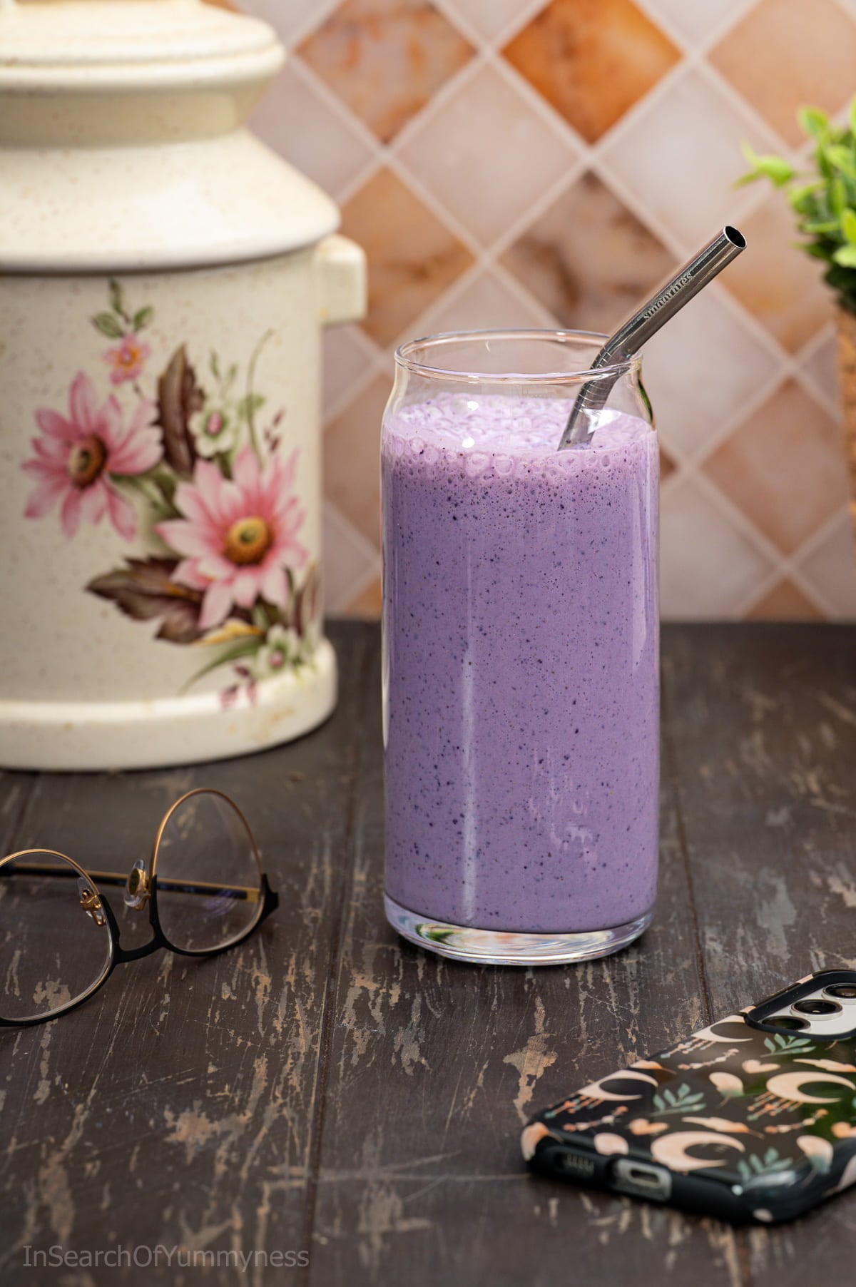 A tall glass filled with purple blueberry oat smoothie, with a metal straw inside. There is a cellphone and a pair of glasses on the table as well.