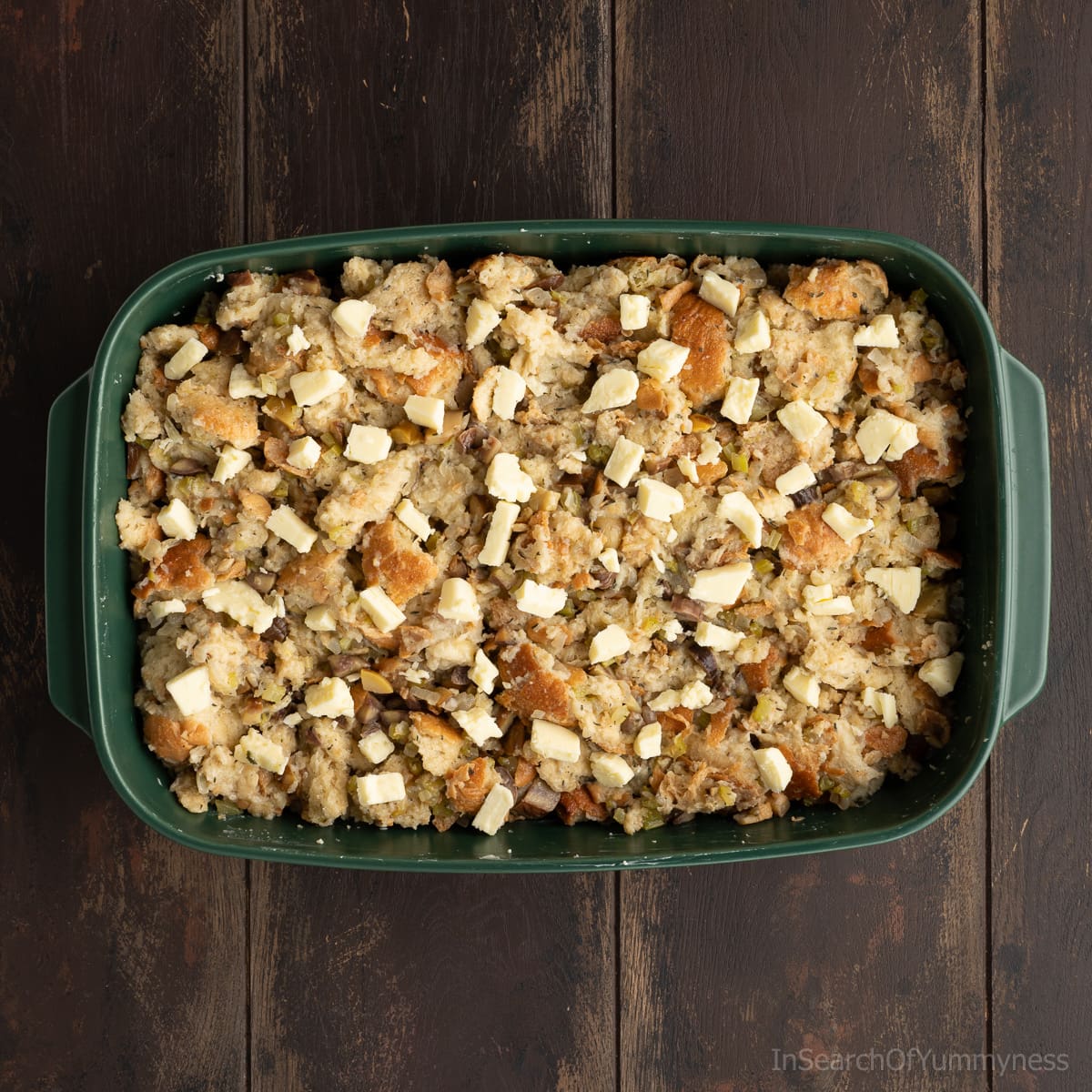 A green baking dish full of the uncooked stuffing mixture, dotted with pieces of cold salted butter.