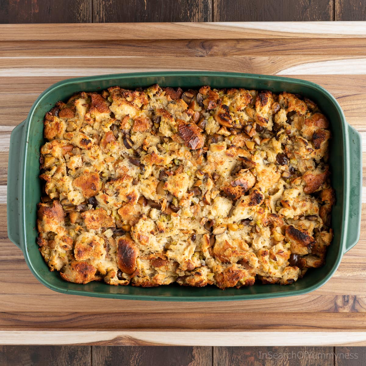 A green dish with baked chestnut stuffing sitting on a cutting board on a dark brown wooden table.