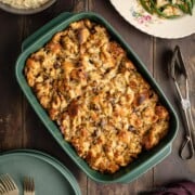 A green baking dish full of freshly baked chestnut stuffing, next to a pair of fancy silver serving tongs, a stack of green plates, and other holiday side dishes.