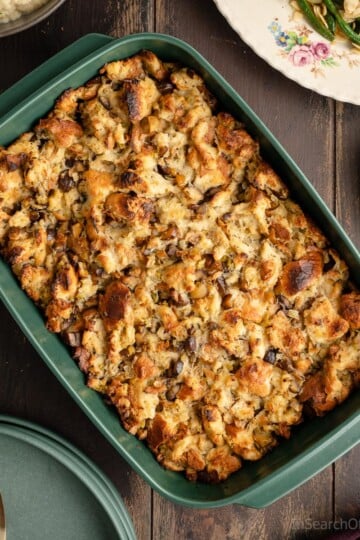 A green baking dish full of freshly baked chestnut stuffing, next to a pair of fancy silver serving tongs, a stack of green plates, and other holiday side dishes.
