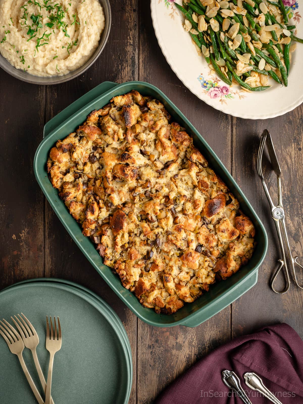 A green Emile Henry baking dish full of freshly baked chestnut stuffing, next to a pair of fancy silver serving tongs, a stack of green plates, and other holiday side dishes.