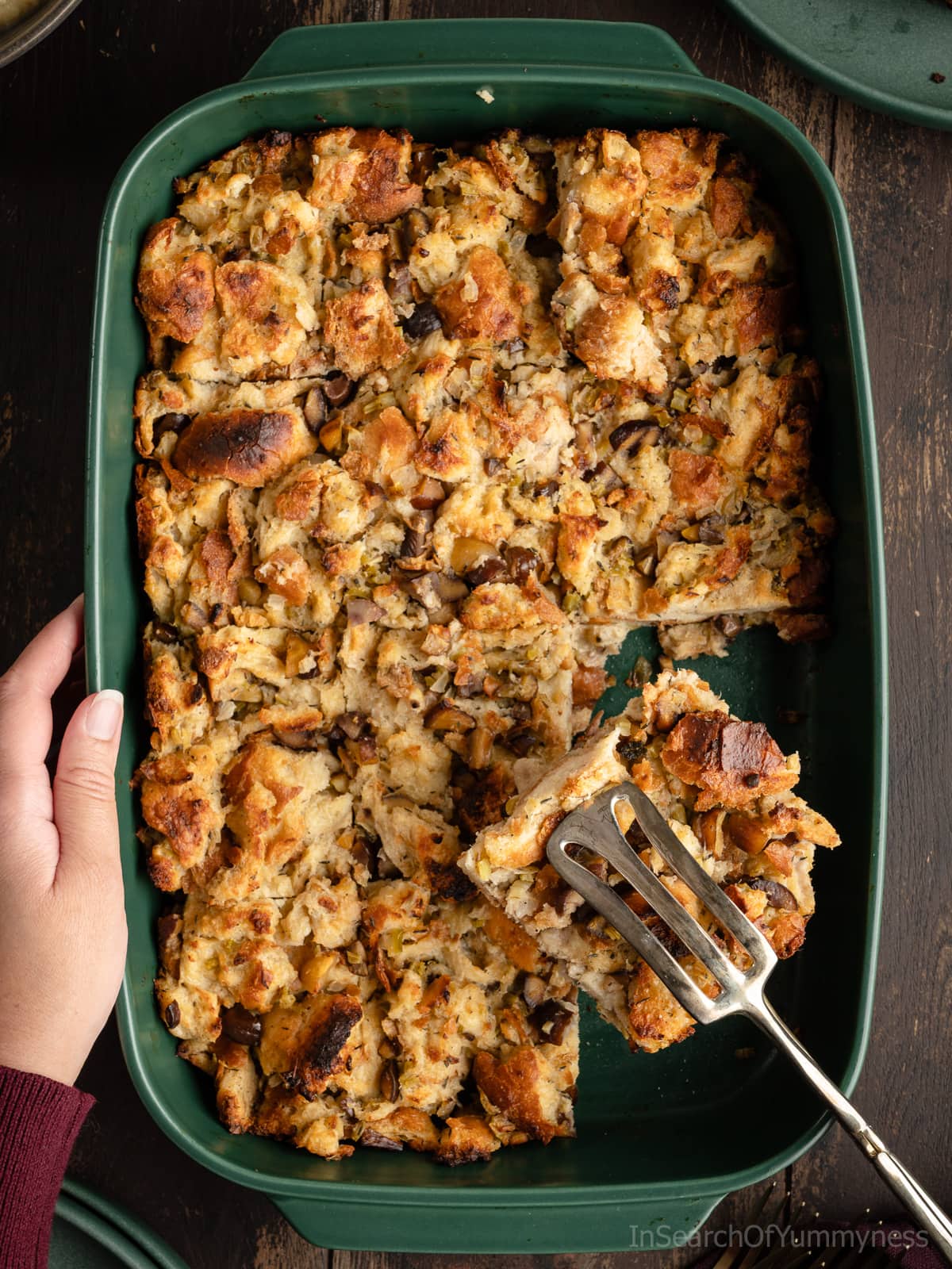 Someone using a pair of fancy serving tongs to serve a slice of baked chestnut stuffing from a green baking dish.