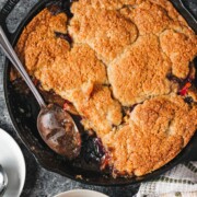 A baked plum cobbler with a buttery biscuit topping in a cast iron pan.