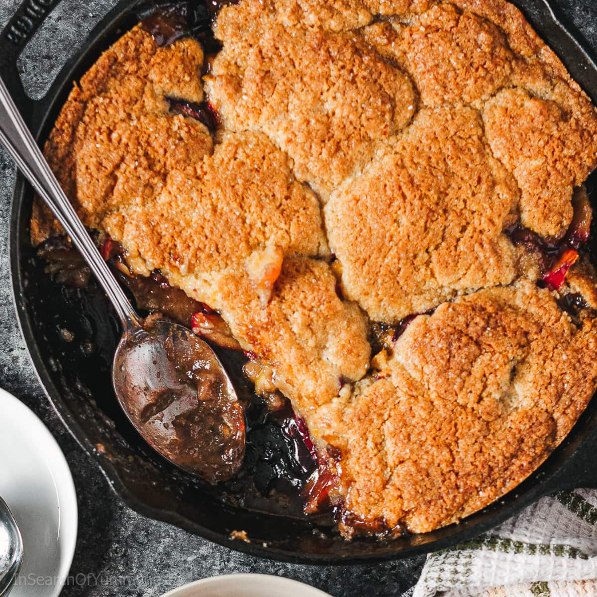 A baked plum cobbler with a buttery biscuit topping in a cast iron pan.