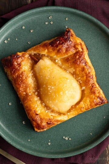 A baked spiced pear in golden brown puff pastry sprinkled with coarse sugar on a green plate, with a purple napkin and gold fork.