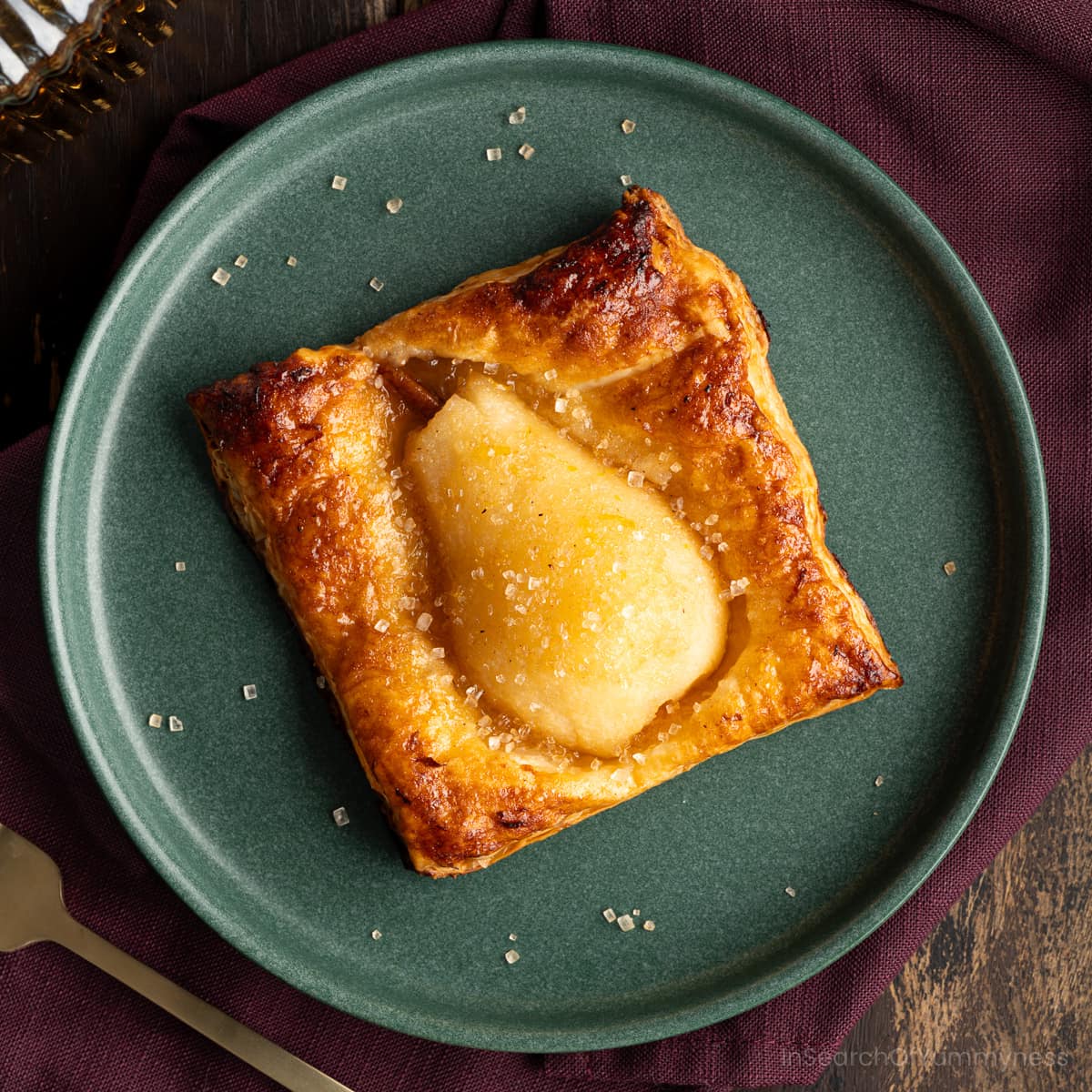 A baked spiced pear in golden brown puff pastry sprinkled with coarse sugar on a green plate, with a purple napkin and gold fork.