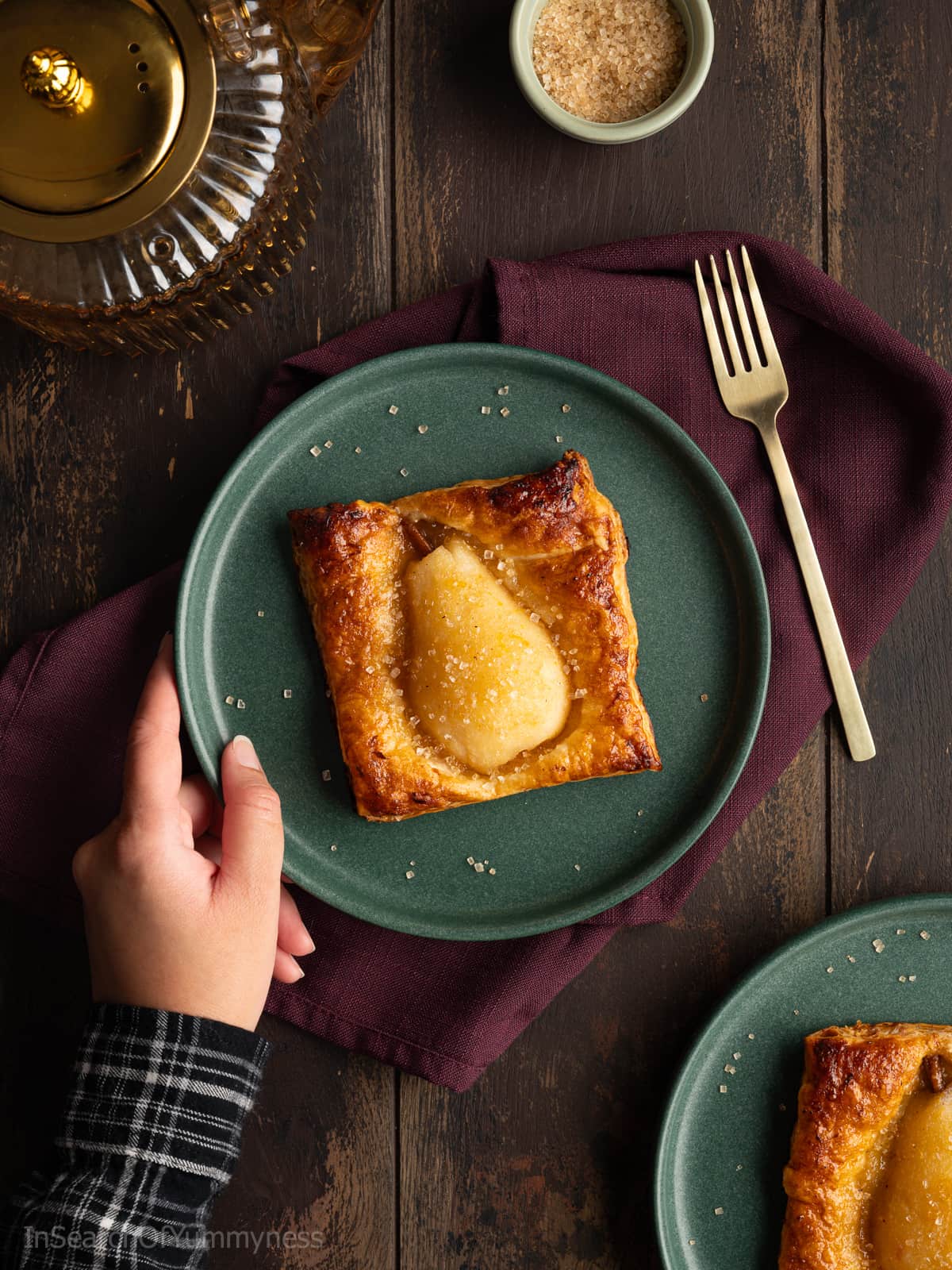 Someone placing a green plate with a baked spiced pear in golden brown puff pastry, sprinkled with coarse sugar, onto a purple napkin.