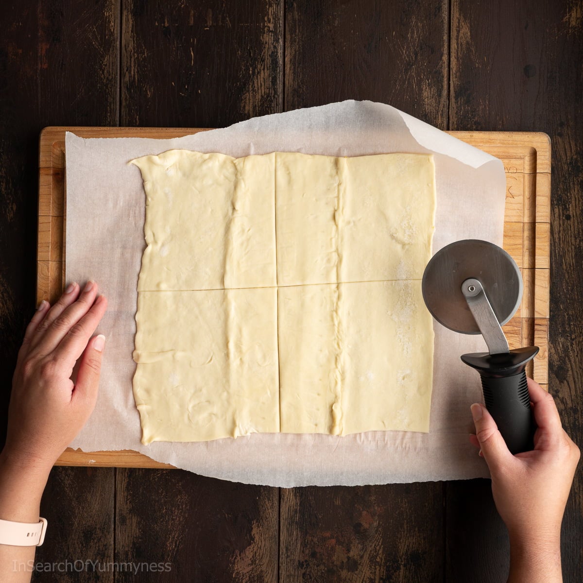 Someone using a pizza cutter to cut a sheet of thawed puff pastry into 4 equal pieces.