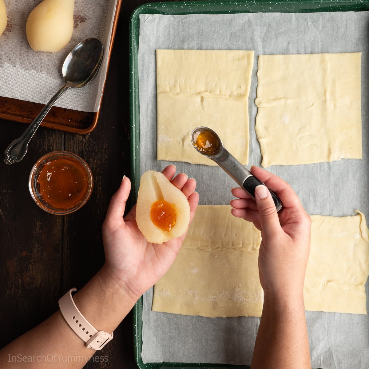 Someone adding a teaspoon of apricot jam to the middle of a canned pear half, over a sheet pan with four pieces of puff pastry.