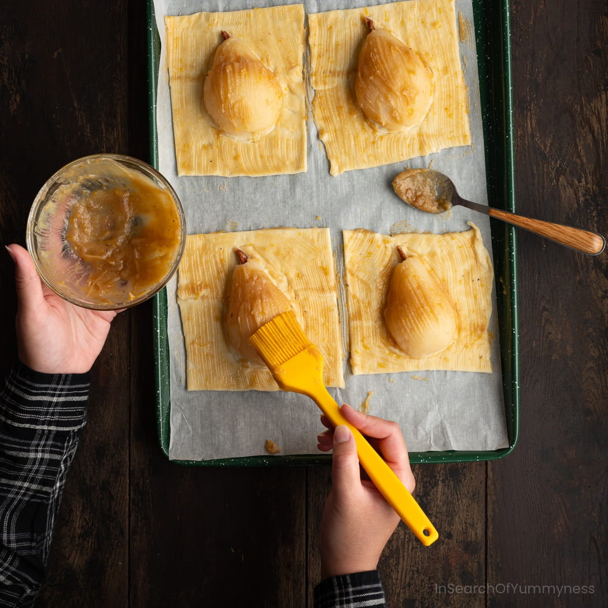 Someone brushing a spiced butter mixture onto four unbaked pears on puff pastry waiting to go into the oven.
