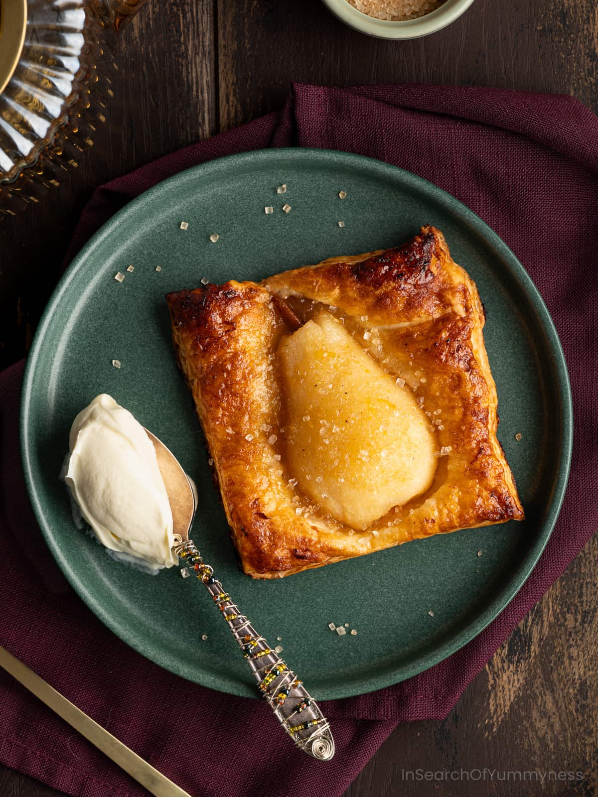 A green plate with a baked spiced pear in golden brown puff pastry, sprinkled with coarse sugar, on a purple napkin. There is a spoon with thick cream next to the patsry.
